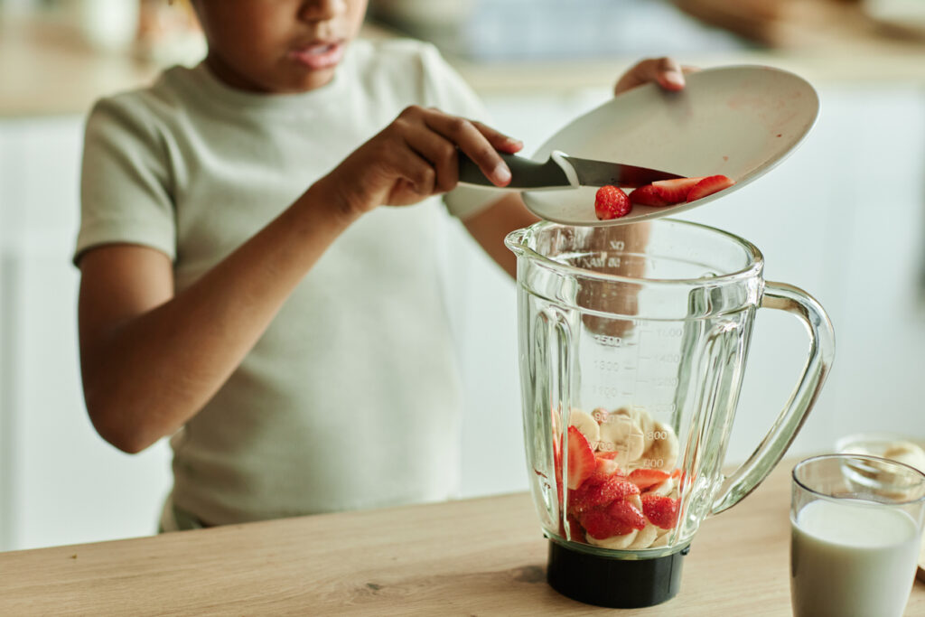 Cropped shot of African American little girl carefully adding strawberries to blender making fruit smoothie with banana for healthy breakfast on kitchen counter