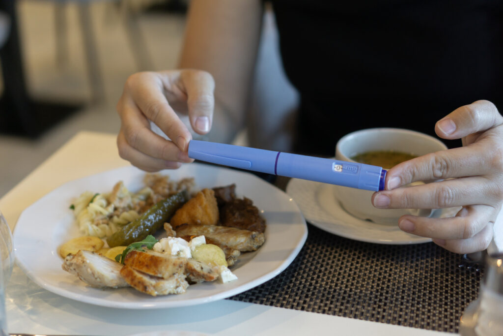 Close-up of hands holding an Ozempic injection pen while sitting at a table with a plate of food and a cup of soup